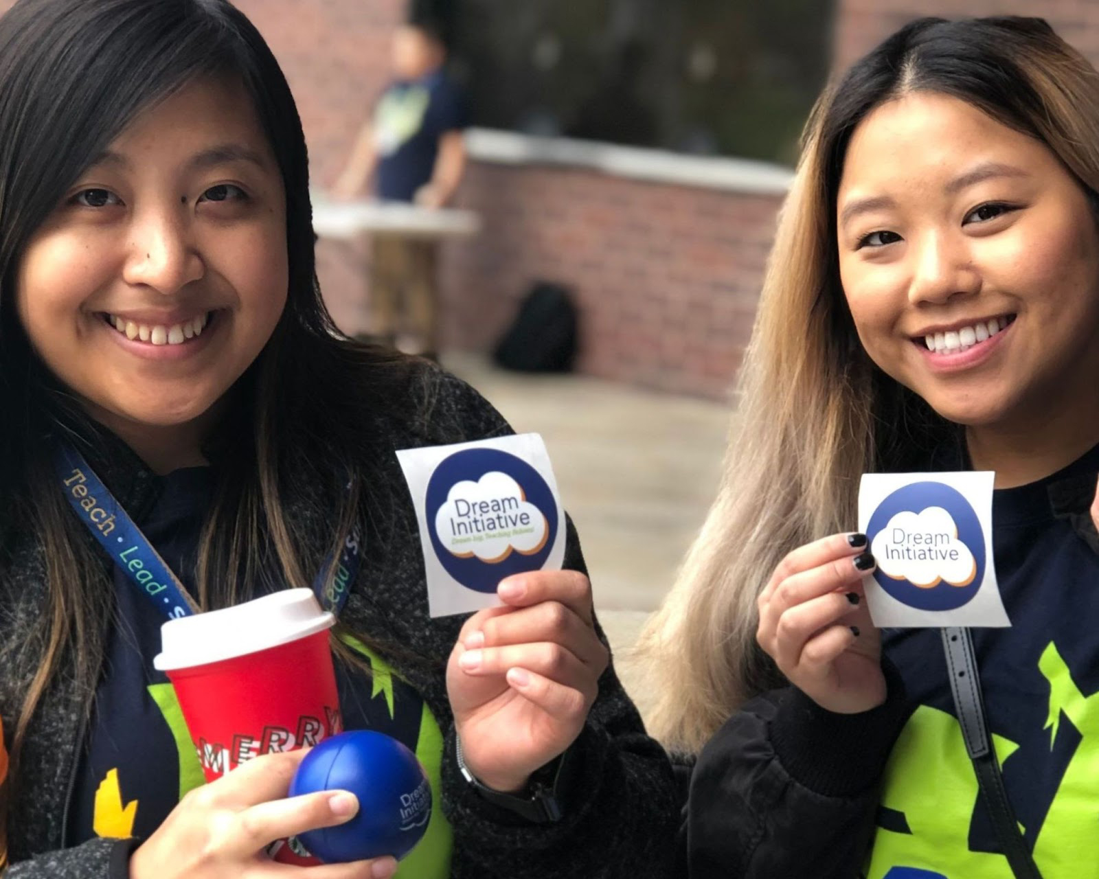 Two smiling Teaching Fellows tutors standing in front of a brick wall holding stickers and a blue stress ball that all say Dream Initiative.