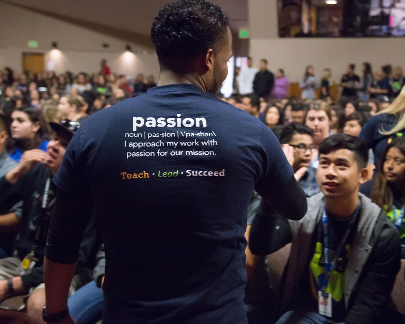 Man in Teaching Fellows shirt with the word PASSION on the back facing an audience of Teaching Fellows tutors in large auditorium.