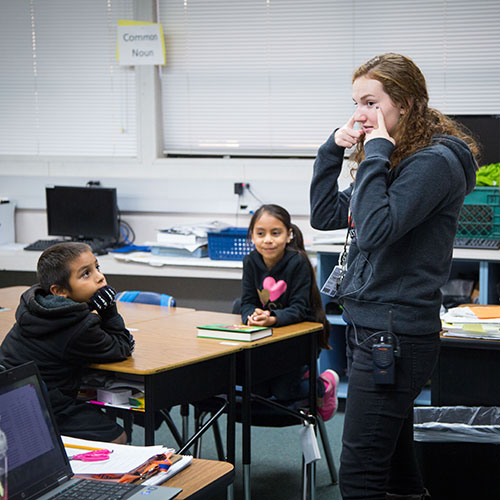 A Teaching Fellow stands at the front of a classroom, pointing to her eyes with both hands as two students look on from their desks.