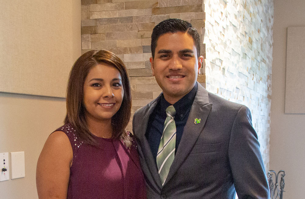 Closeup of a woman in a purple dress and a man in a gray suit and tie standing close together in front of a stacked stone wall and both people smiling at the camera.