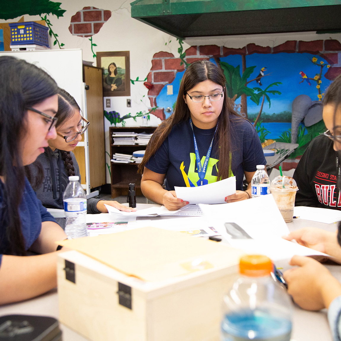 A group of 4 people all wearing reading glasses, sitting at a table in a classroom looking at papers.