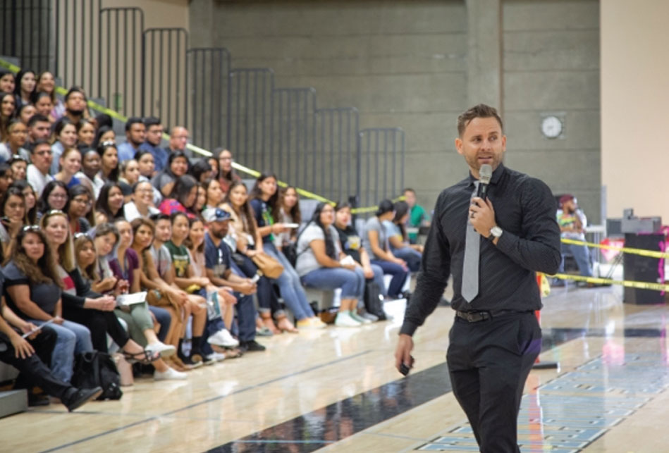 Man in black shirt and pants with grey neck tie talking on microphone in front of a gymnasium full of Teaching Fellows tutors sitting in the bleachers.